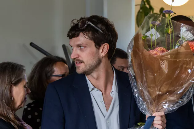 A person with a black jacket and a white shirt. The person is holding flowers.